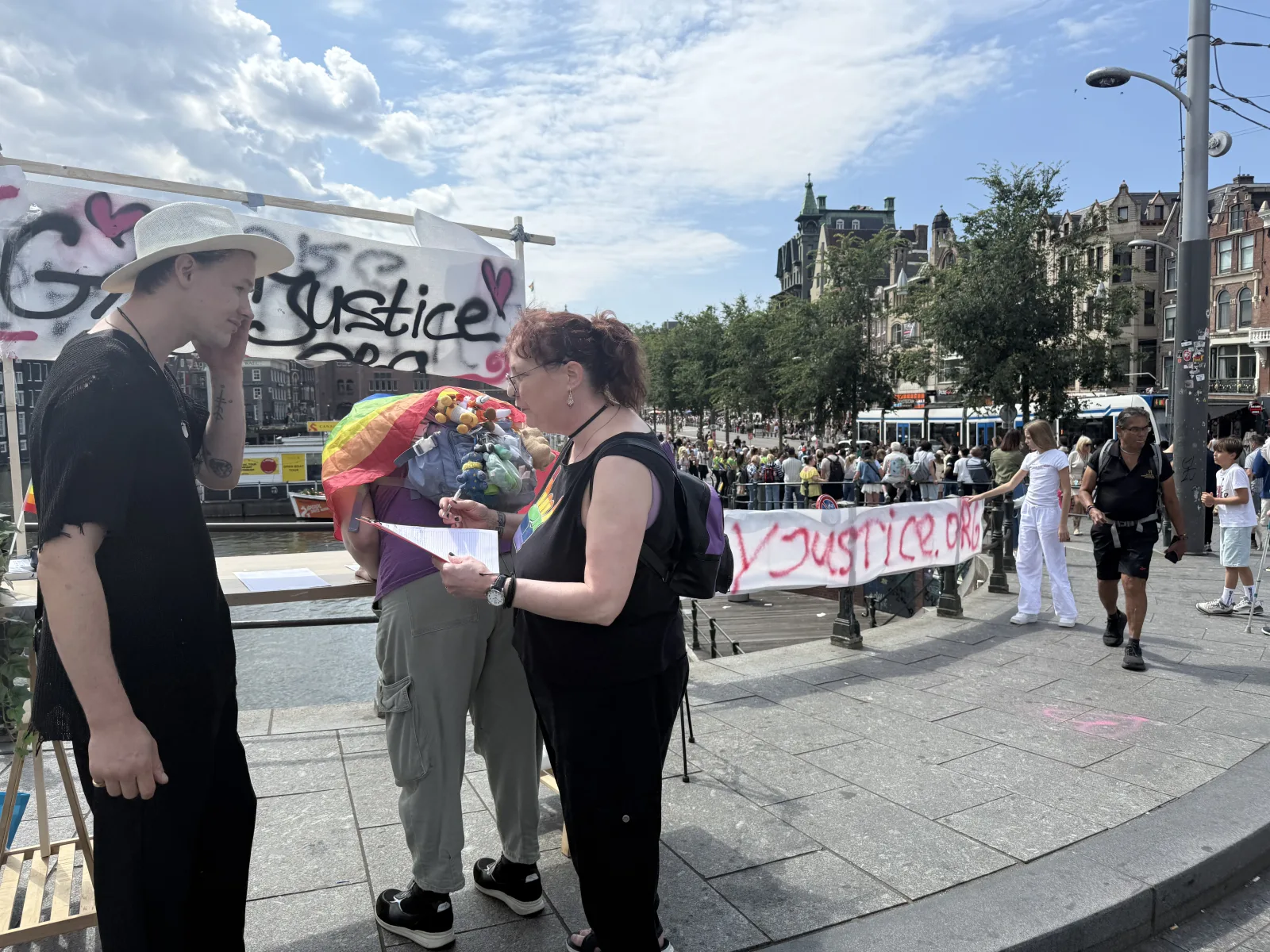 A petition table and WorldPride crowd behind the GayJustice campaign display in Amsterdam.
